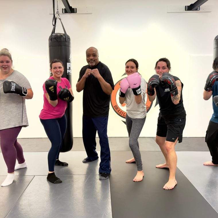 Students wearing boxing gloves pose with fists raised alongside their instructor on the gym mats.