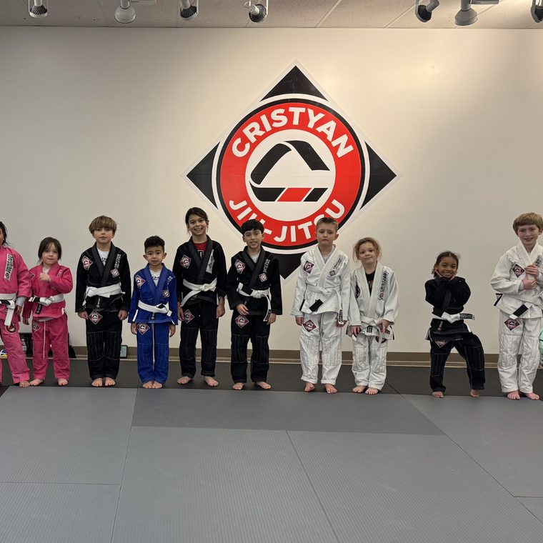 A line of young children in colored gis standing side by side on the training mats at Cristyan Jiu-Jitsu.