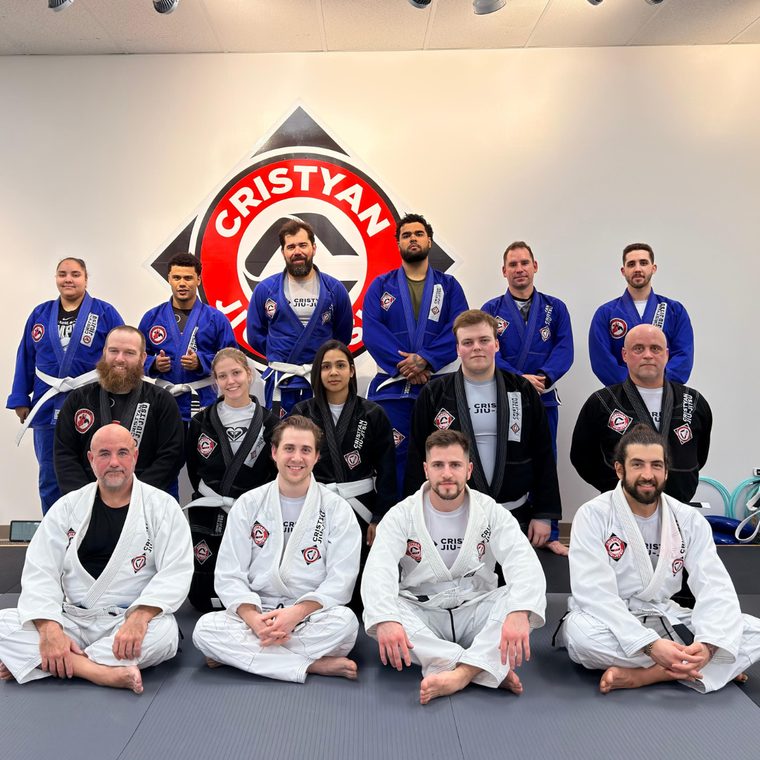 Adult students in white, black and blue gis posed in front of the Cristyan Jiu-Jitsu wall logo.