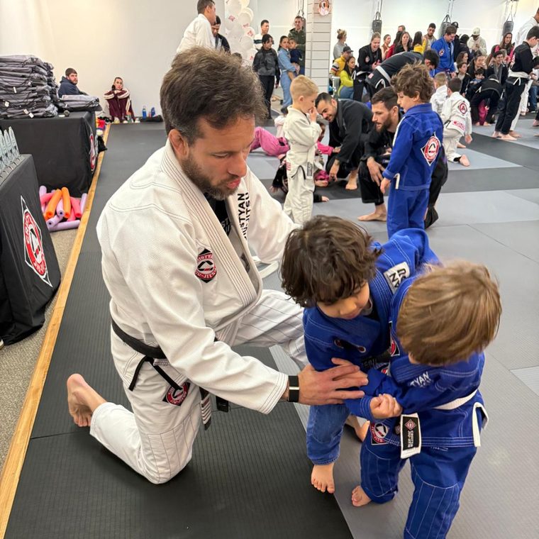 A black belt instructor kneeling on the mats to coach two young children wearing blue gis during a kids' jiu-jitsu class.